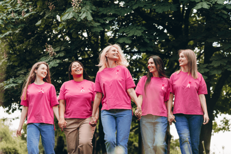 Cinque donne sorridenti camminano mano nella mano in un parco, indossando magliette rosa con il nastro simbolo della prevenzione del tumore al seno, durante un evento di sensibilizzazione per l’Ottobre Rosa.
