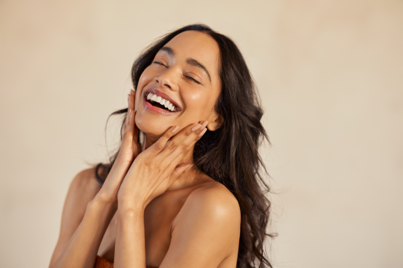Donna sorridente che si tocca il viso, immagine associata alla cura della pelle dopo l’esposizione al sole e all’importanza dell’idratazione.