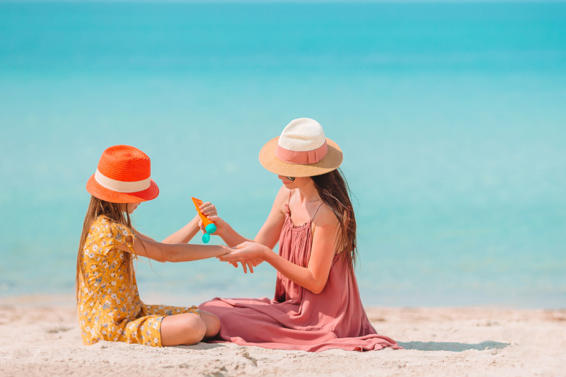 Due bambine sedute sulla spiaggia mentre un’adulta applica la protezione solare sul braccio di una di loro, con mare sullo sfondo.